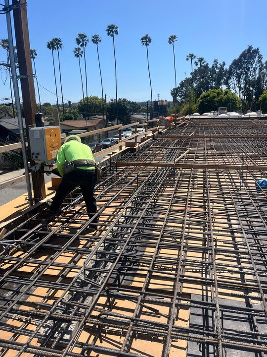 Construction worker installing rebar on a building foundation with palm trees and blue sky in the background.