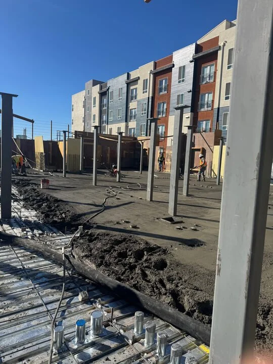 Construction site with a multi-story apartment building in the background and ongoing concrete work in the foreground.