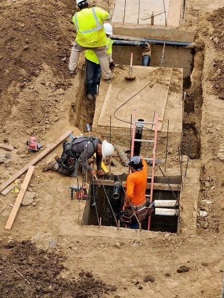 Three construction workers, wearing safety helmets and vests, are working in an excavation site. They are installing or repairing underground pipes or utilities within a deep, rectangular trench.