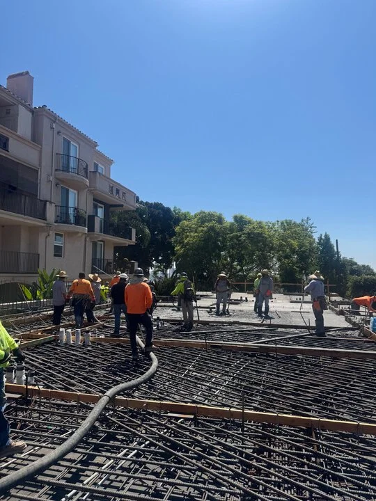 Construction workers pouring concrete on a building site with rebar reinforcement, next to a residential building and trees, under a clear blue sky.
