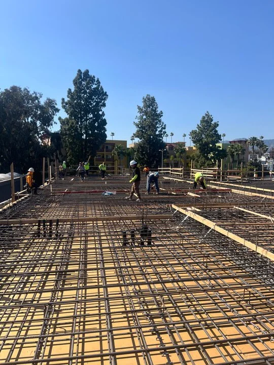 Construction workers working on rebar reinforcement for a building's concrete floor or foundation, outdoors on a sunny day.