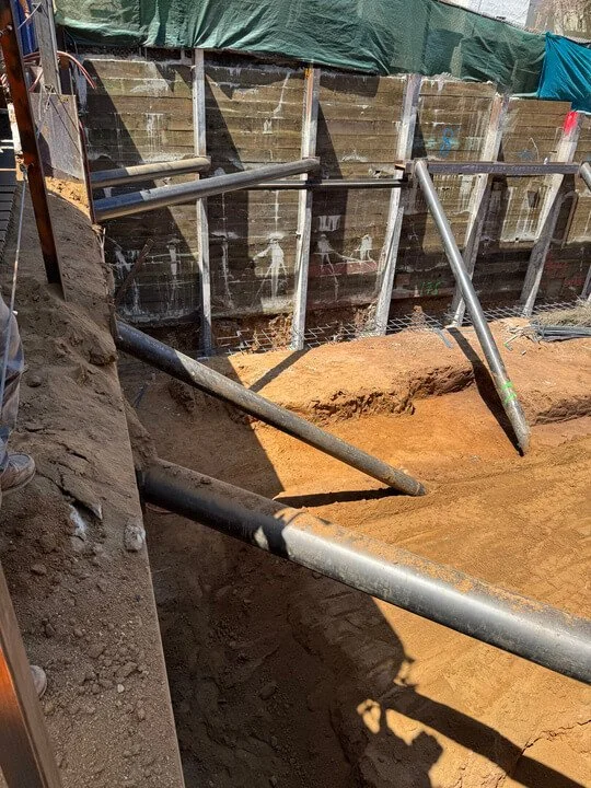 Construction site with metal poles and excavated dirt, behind a wooden fence with green and blue tarps.