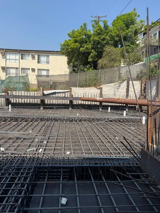 Construction site with steel rebar grid for concrete slab, surrounded by a fence and nearby residential buildings and trees.