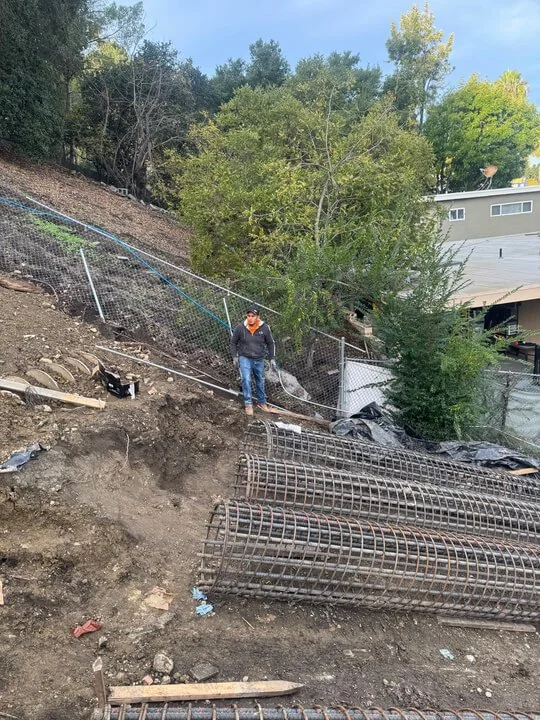 A construction site on a hillside with a man standing near steel reinforcement pipes, a chain-link fence, and trees in the background.