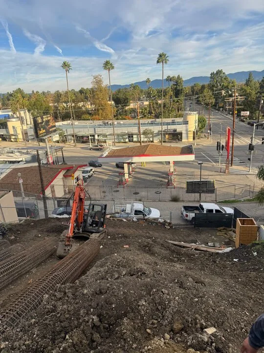 View from a hill showing a city street with palm trees, parked cars, a gas station, and mountains in the background under a partly cloudy sky.
