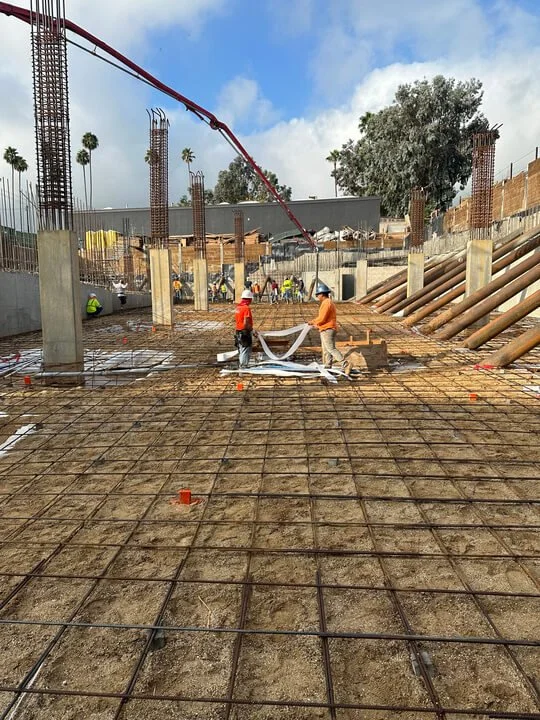 Construction workers pouring and spreading concrete on a foundation with rebar grid, building supports, and equipment in the background.