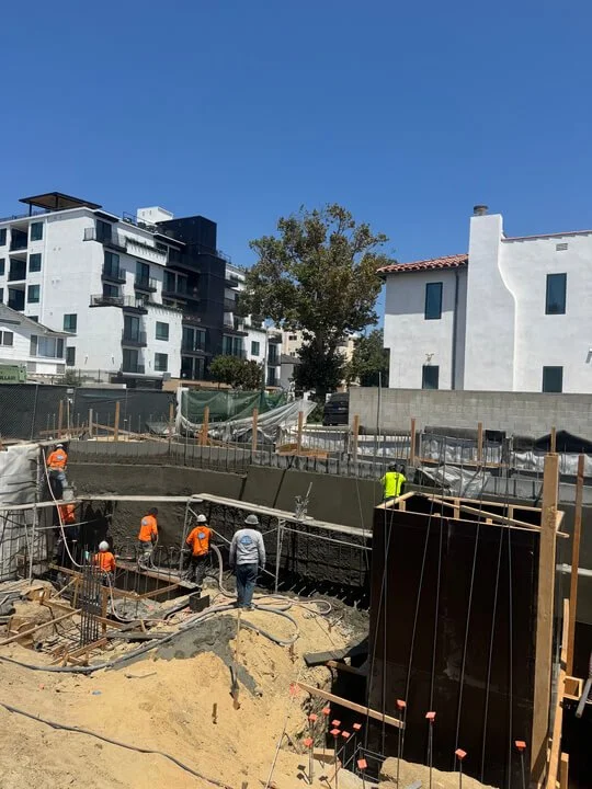 Construction workers working on a building foundation at a construction site, with some workers wearing orange vests and helmets, surrounded by construction materials and equipment, with apartment buildings and trees in the background.
