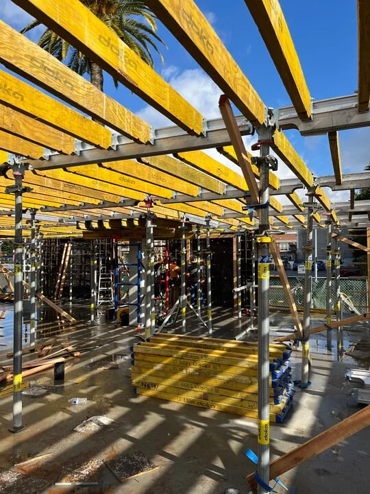 Construction site with scaffolding and yellow safety beams, palm trees and a blue sky in the background.