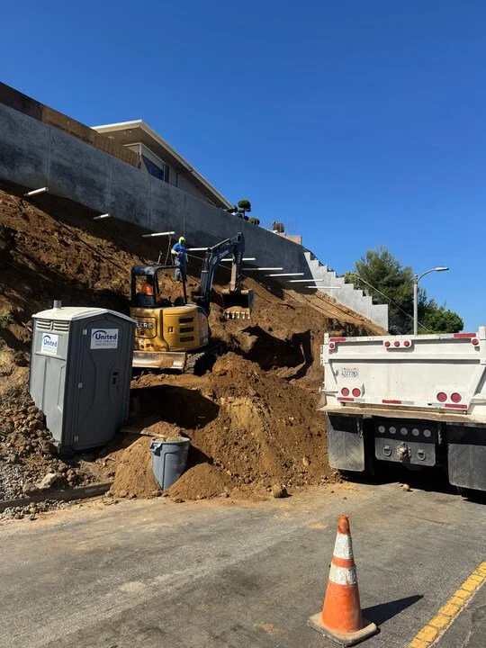 Excavator digging into the hillside near a building, with a portable toilet and a construction truck nearby, and an orange traffic cone in the street.
