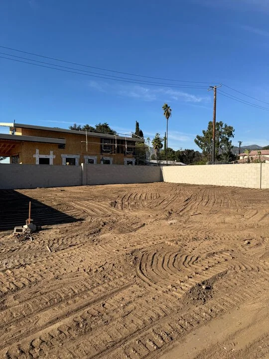 A cleared construction site with dirt ground and tire tracks, surrounded by a concrete wall, with a two-story building and trees in the background under a clear blue sky.