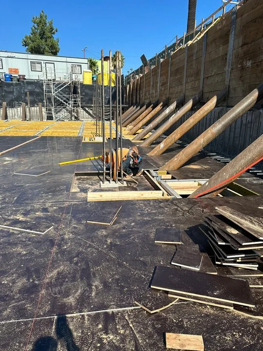 Construction site with worker installing large black tiles on the floor, wooden supports, and a wooden retaining wall at the back.