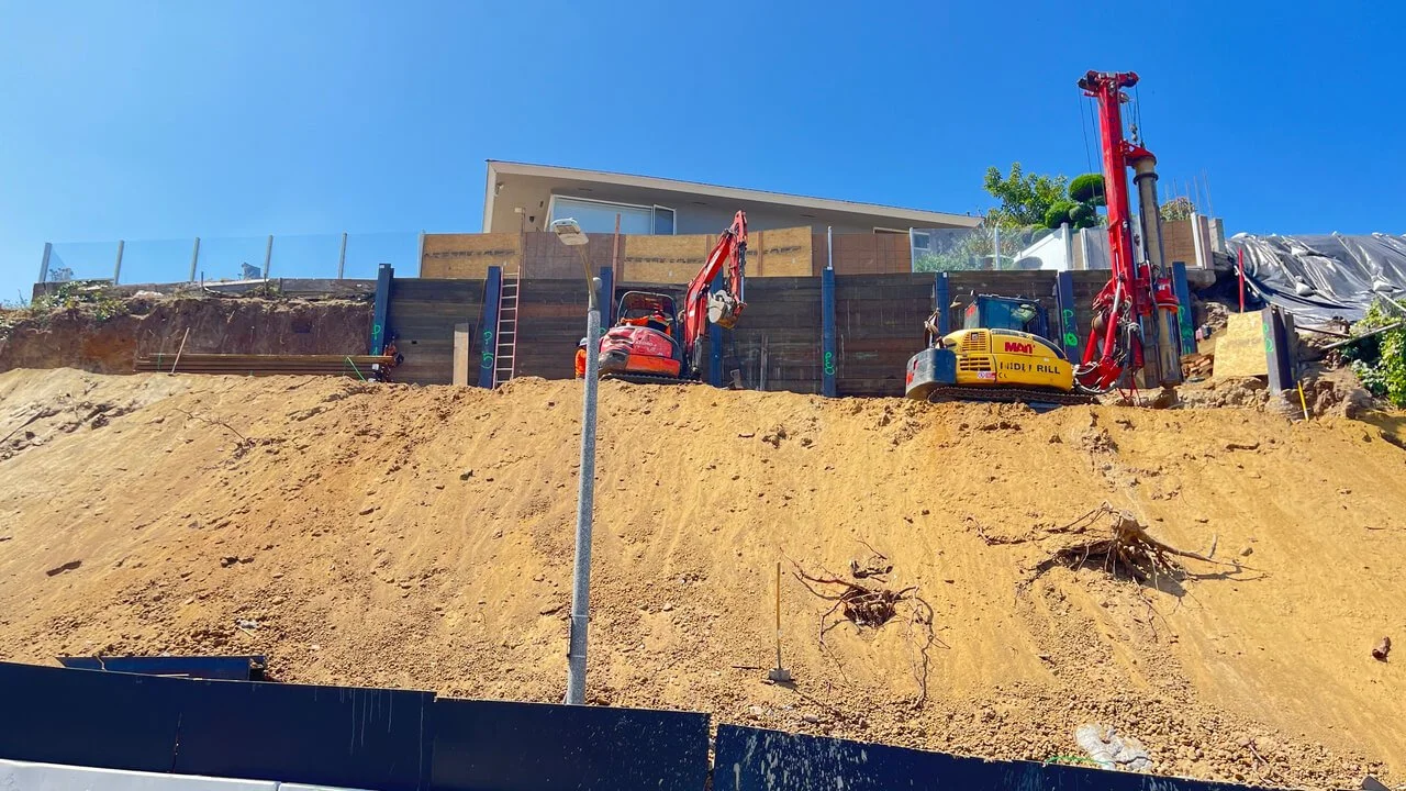 Construction site on a hillside with excavators and a retaining wall, with a modern house on top under a clear blue sky.
