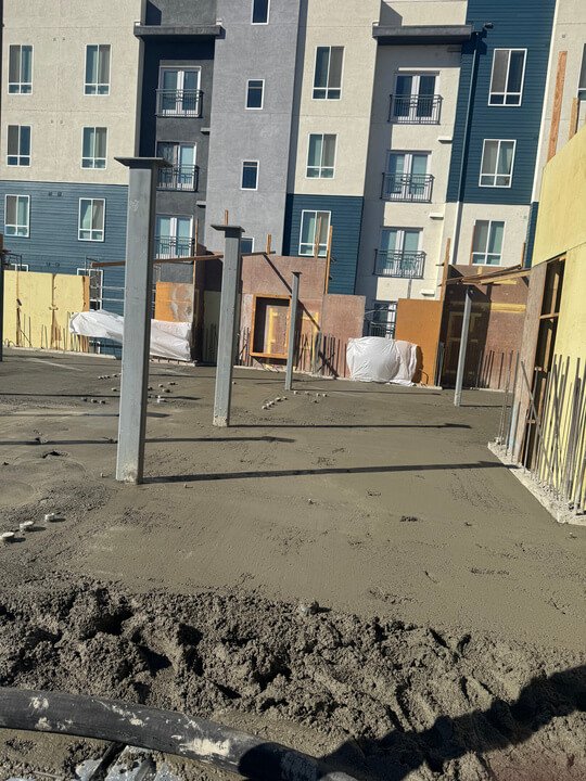 Construction site with steel posts, concrete foundation, and urban apartment buildings in the background.