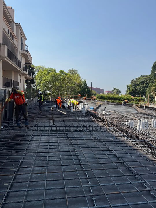 Construction workers installing rebar on a building's rooftop, with a cityscape and trees in the background under a clear blue sky.