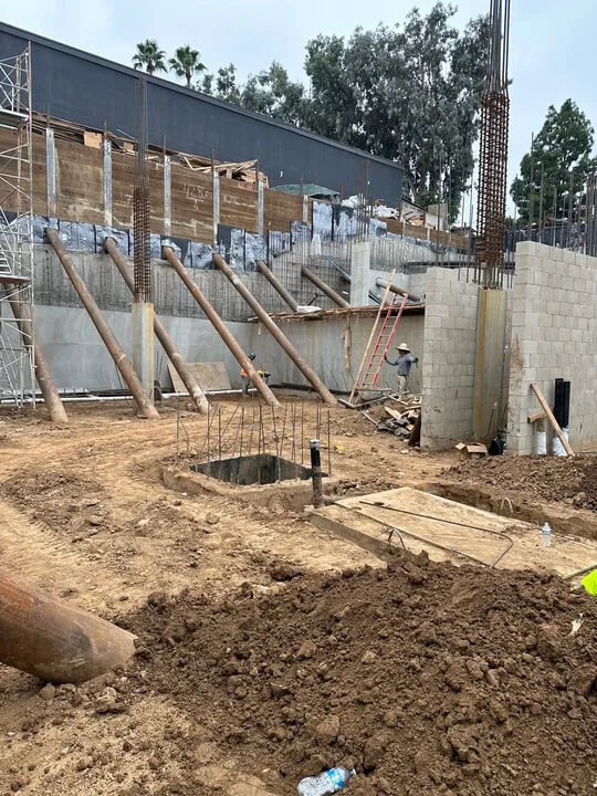 Construction site with concrete wall being built, supported by wooden braces, with building materials and workers present.