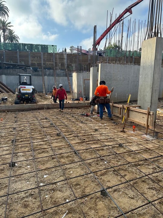 Construction site with workers installing rebar for a concrete slab, with concrete walls and heavy machinery in the background.