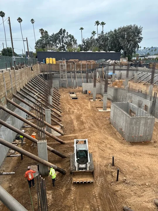 Construction site with excavation, concrete walls, steel reinforcements, and workers operating machinery and tools.