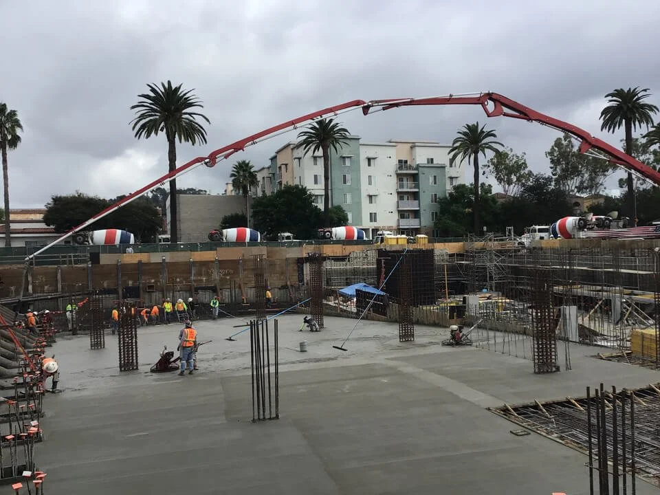 Construction workers pouring and smoothing concrete on a building site with rebar and scaffolding, palm trees in the background, and concrete mixer trucks passing by.