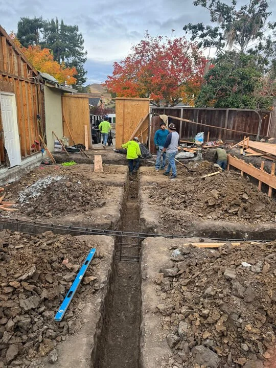 Construction workers excavating and preparing trenches, with some installing rebar, for a building foundation in a backyard, with trees showing fall colors in the background.