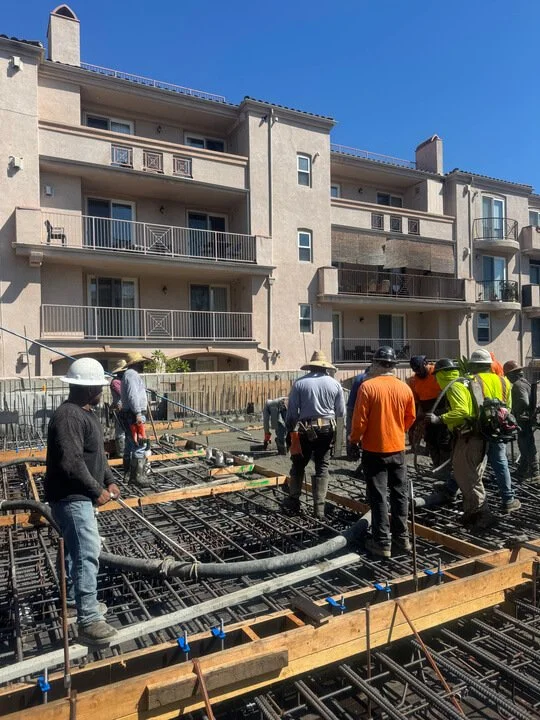 Construction workers on a building site pouring concrete on a rebar framework in front of a multi-story apartment building.