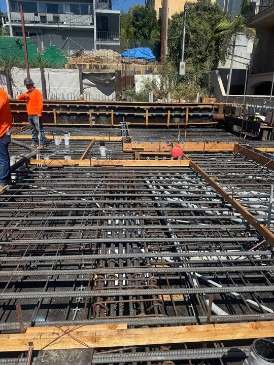 Construction workers on a building site, installing steel reinforcement bars for a concrete floor, with wooden forms and piping visible.