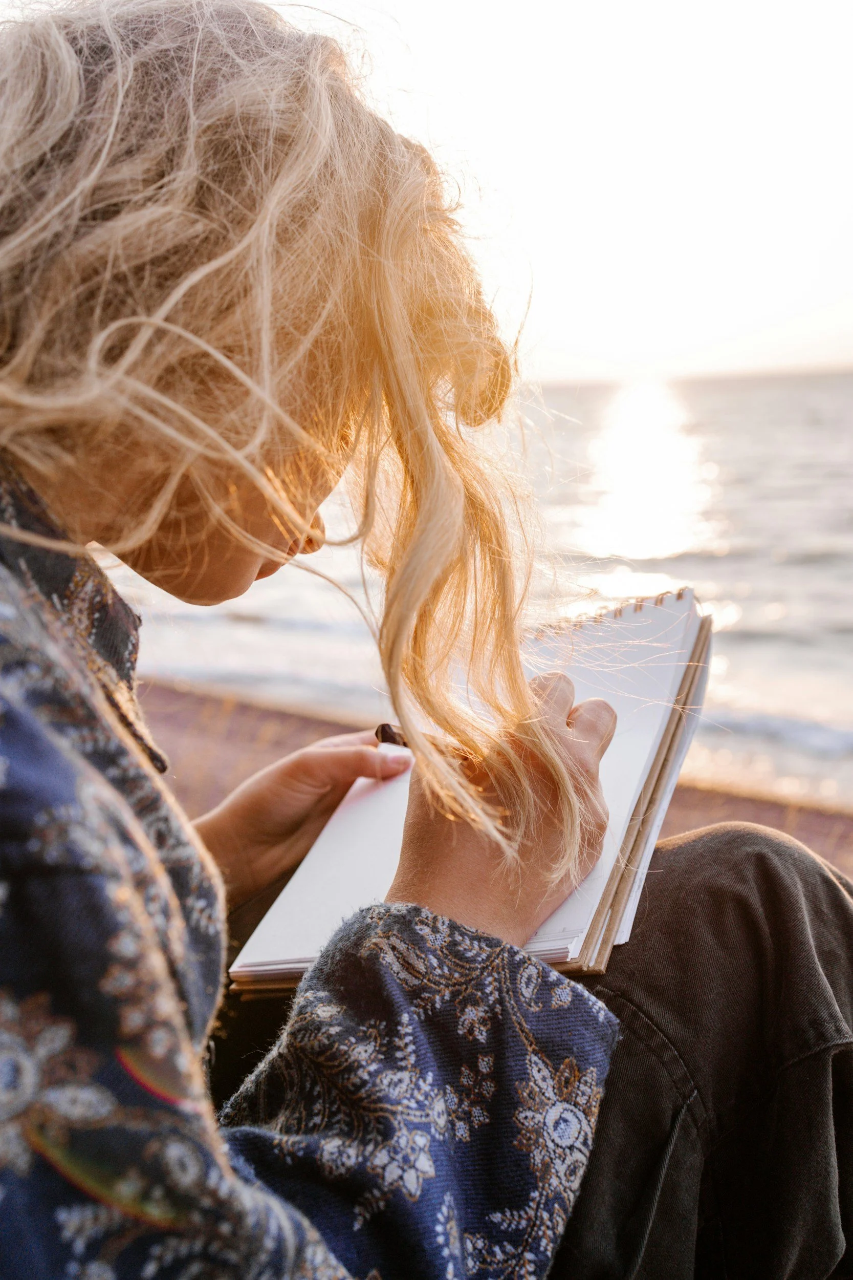 Young woman with blonde curly hair sitting on beach at sunset, writing or drawing in a small notebook.