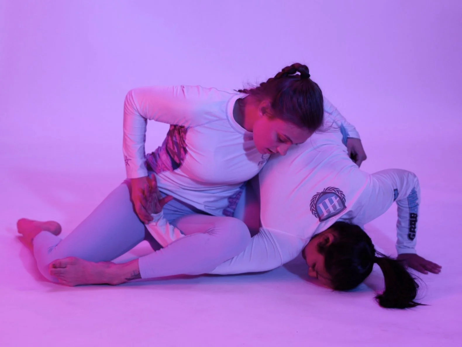 Two women practicing Brazilian Jiu-Jitsu on a pink and purple-lit floor, wearing white Gis, engaged in a grappling position.