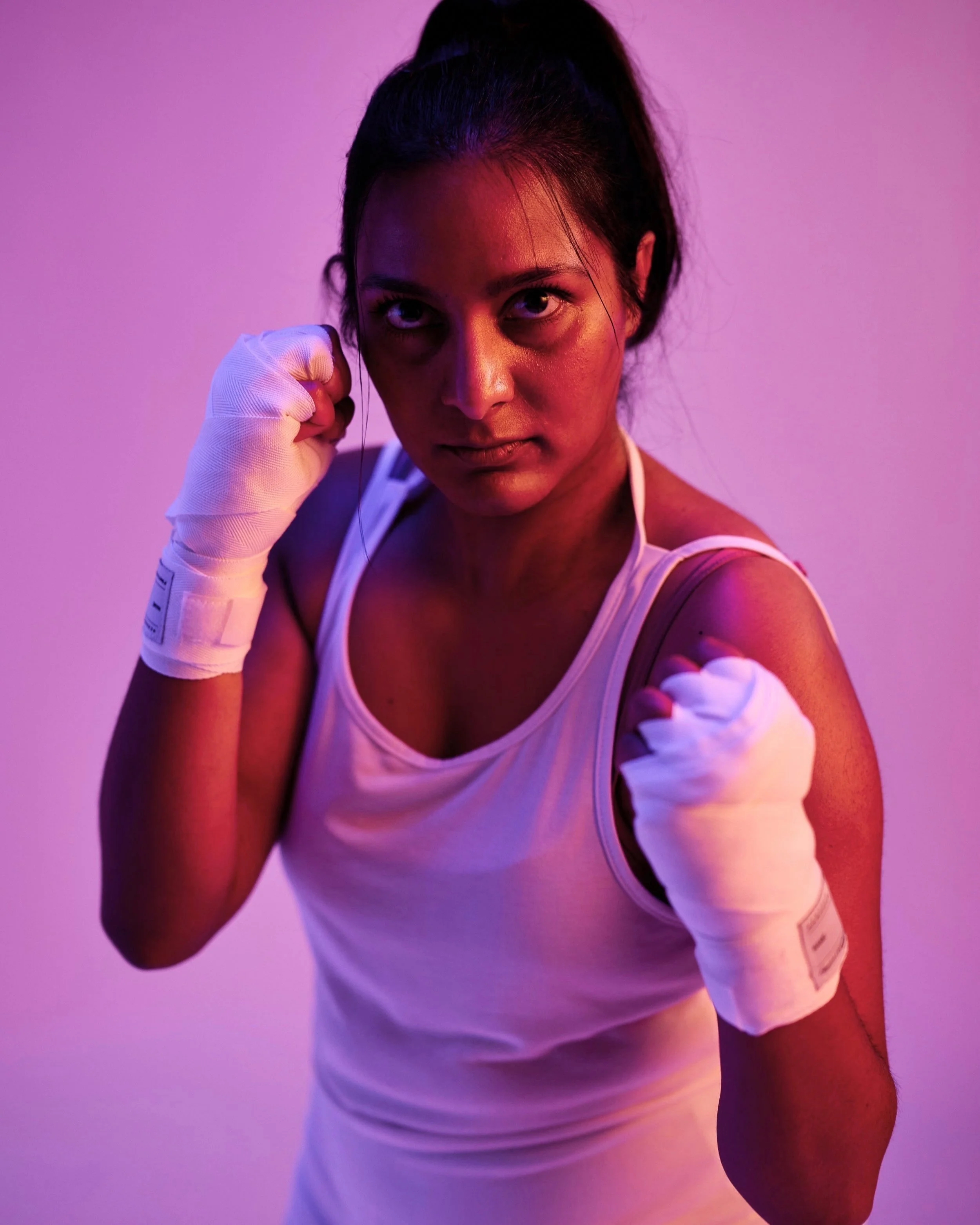 A woman in a white tank top, with her fists up in a boxing stance, wearing white hand wraps, intense expression, against a colorful purple-pink background with dramatic lighting.