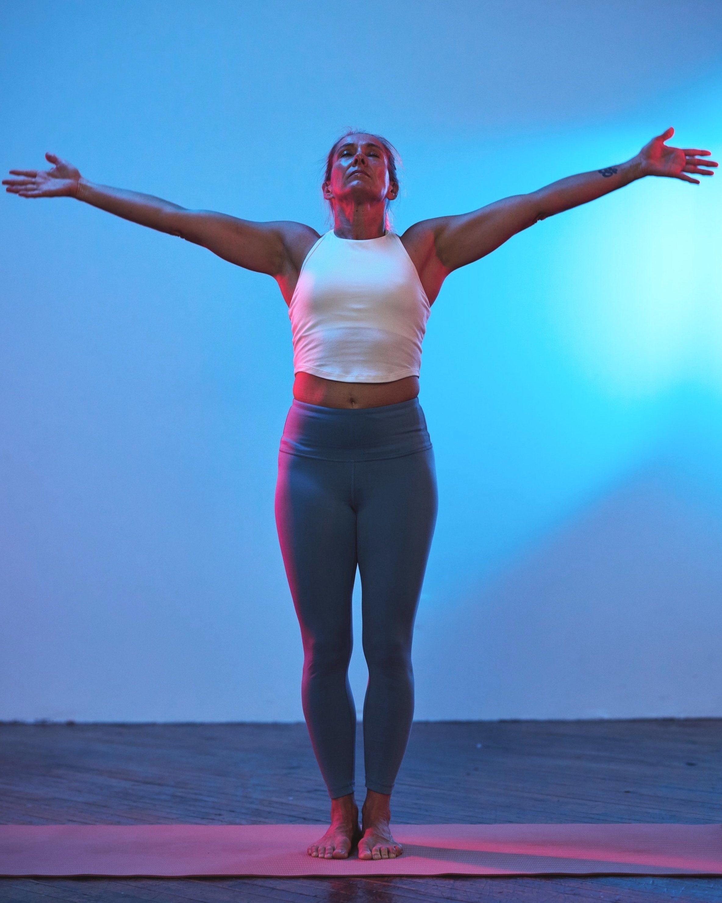 A woman practicing yoga indoors, standing on a yoga mat with arms extended outwards and eyes closed.