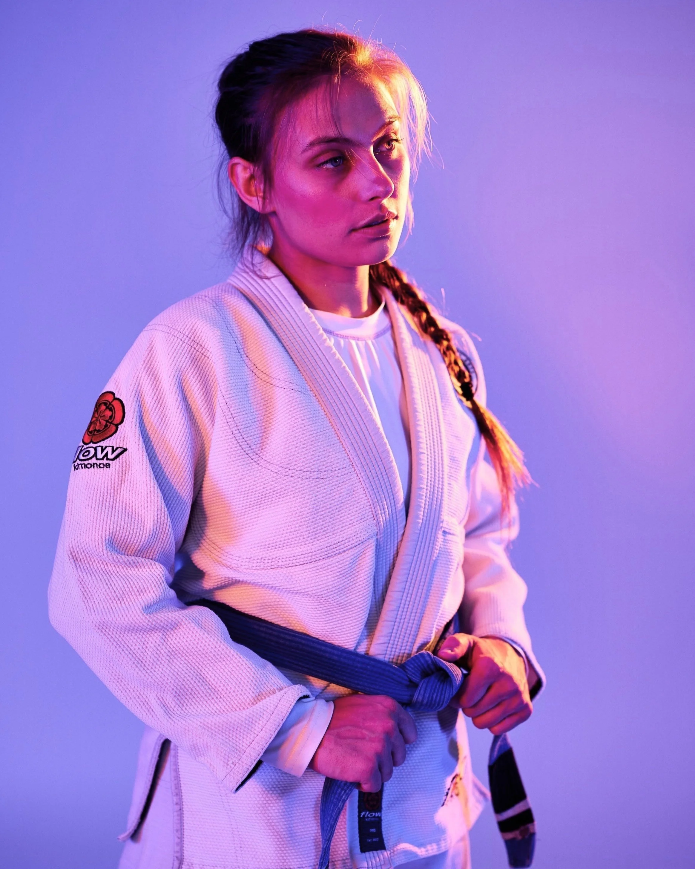 A woman wearing a white Brazilian Jiu-Jitsu gi with a gray belt, standing against a purple background illuminated with colorful lighting.