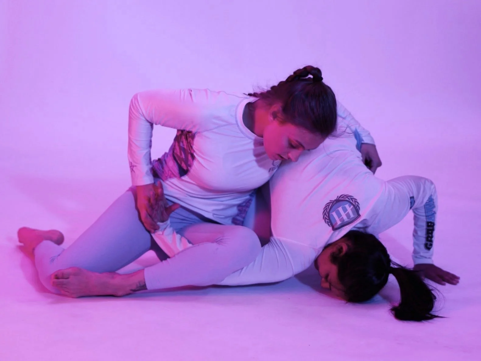 Two women practicing Brazilian Jiu-Jitsu on a purple-lit floor, wearing white gis with matching patches, engaged in a grappling move.