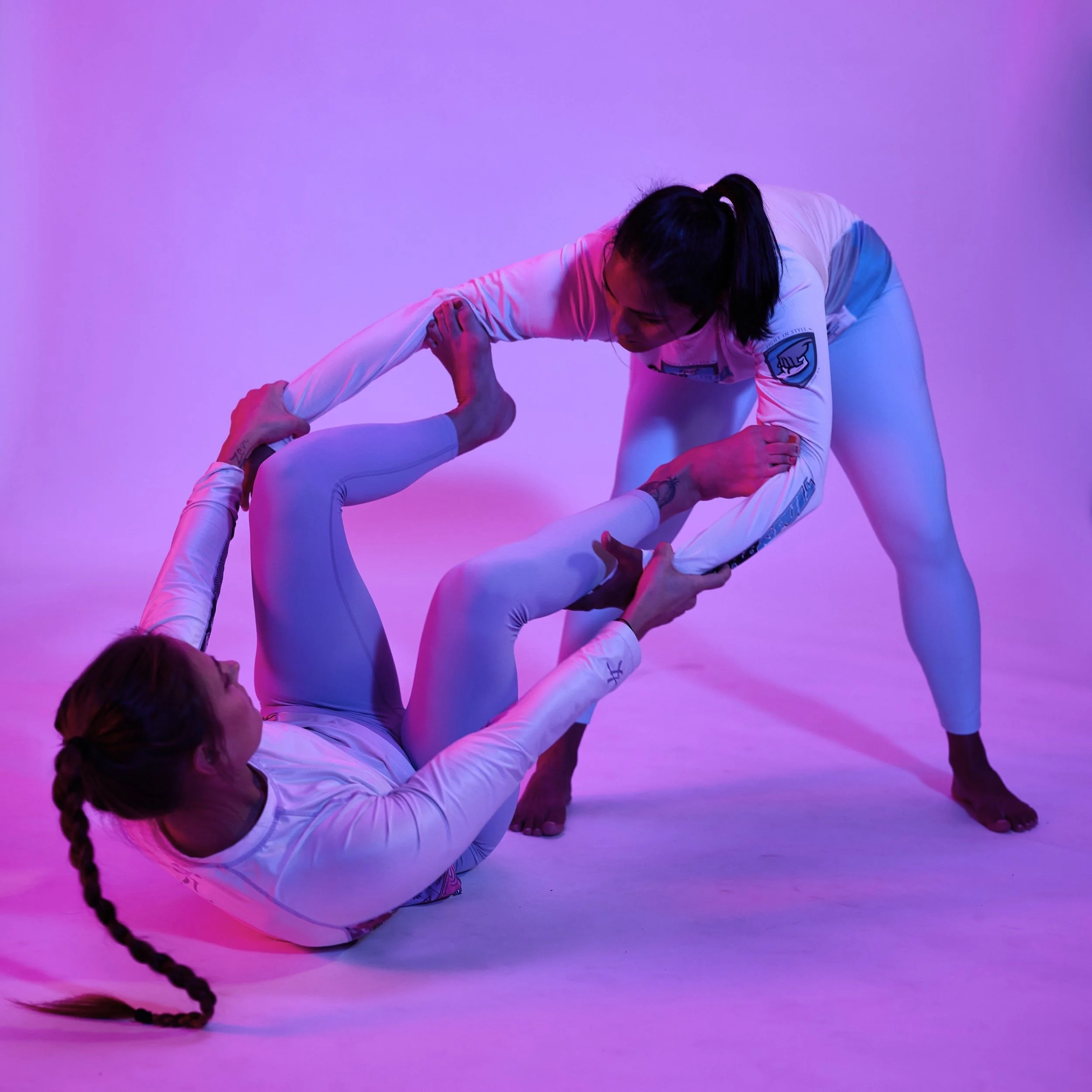 Two women practicing Brazilian Jiu-Jitsu in a studio with purple lighting, one woman on the floor holding the other's leg while the other woman supports herself with her hands on the floor.