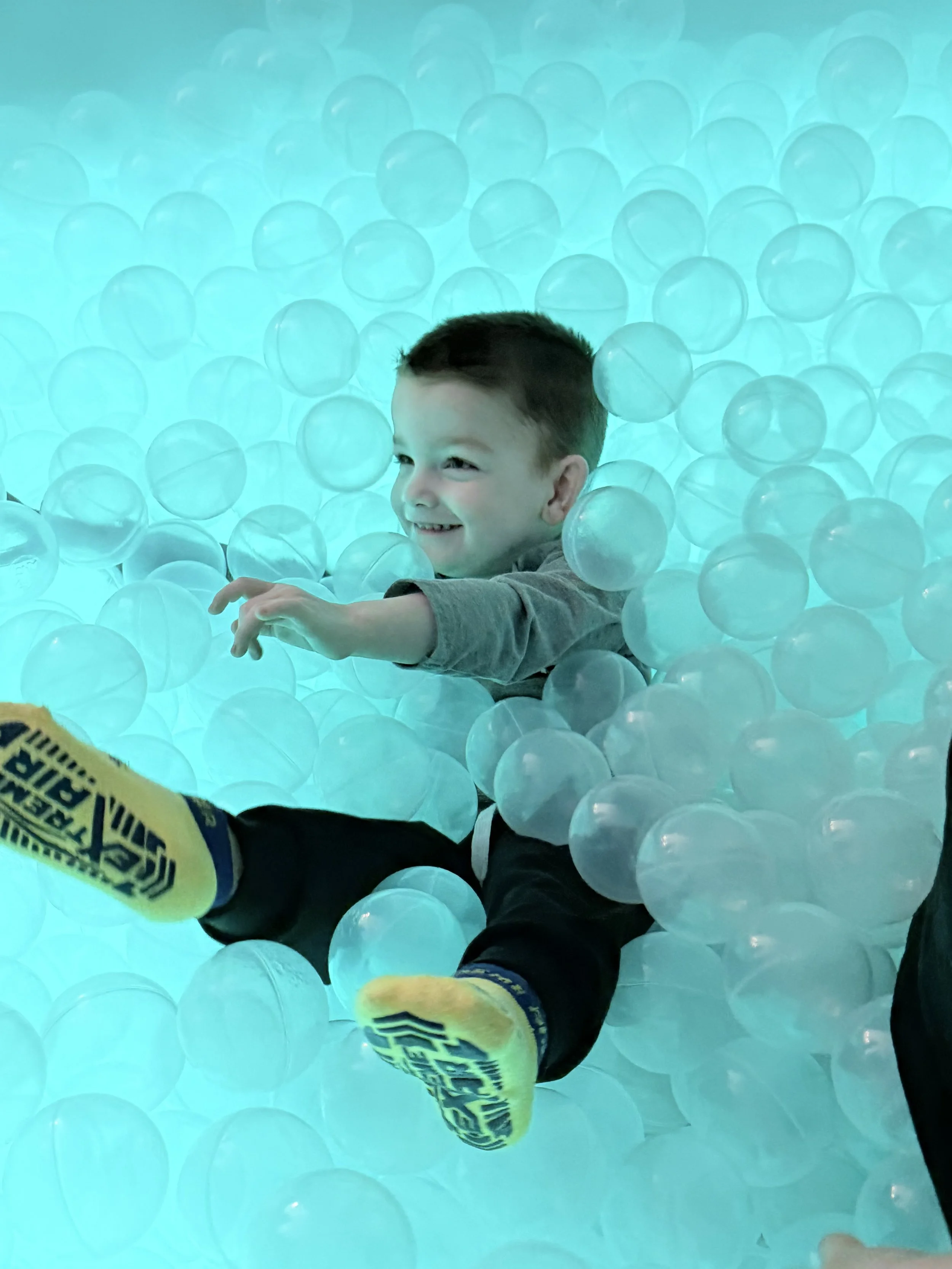 A young boy smiling and playing in a ball pit filled with blue translucent balls.