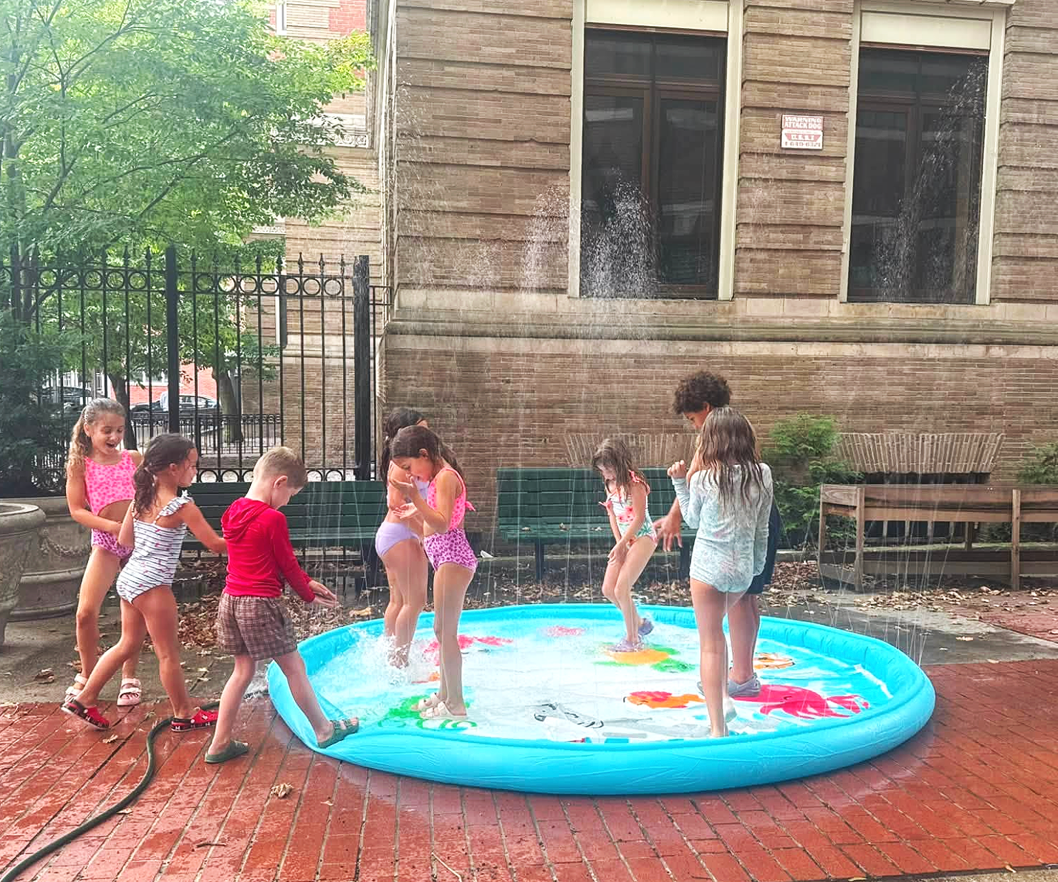 Children playing in a small inflatable pool on a brick patio with a brick building and trees in the background.