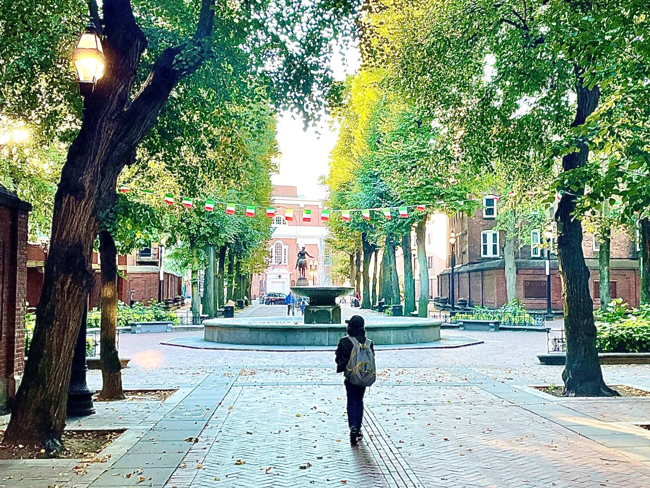 Park with large trees, a fountain, and a brick building in the background. Two people walk on a paved path, one wearing a backpack. Decorations of red, white, and green flags hang across the trees.