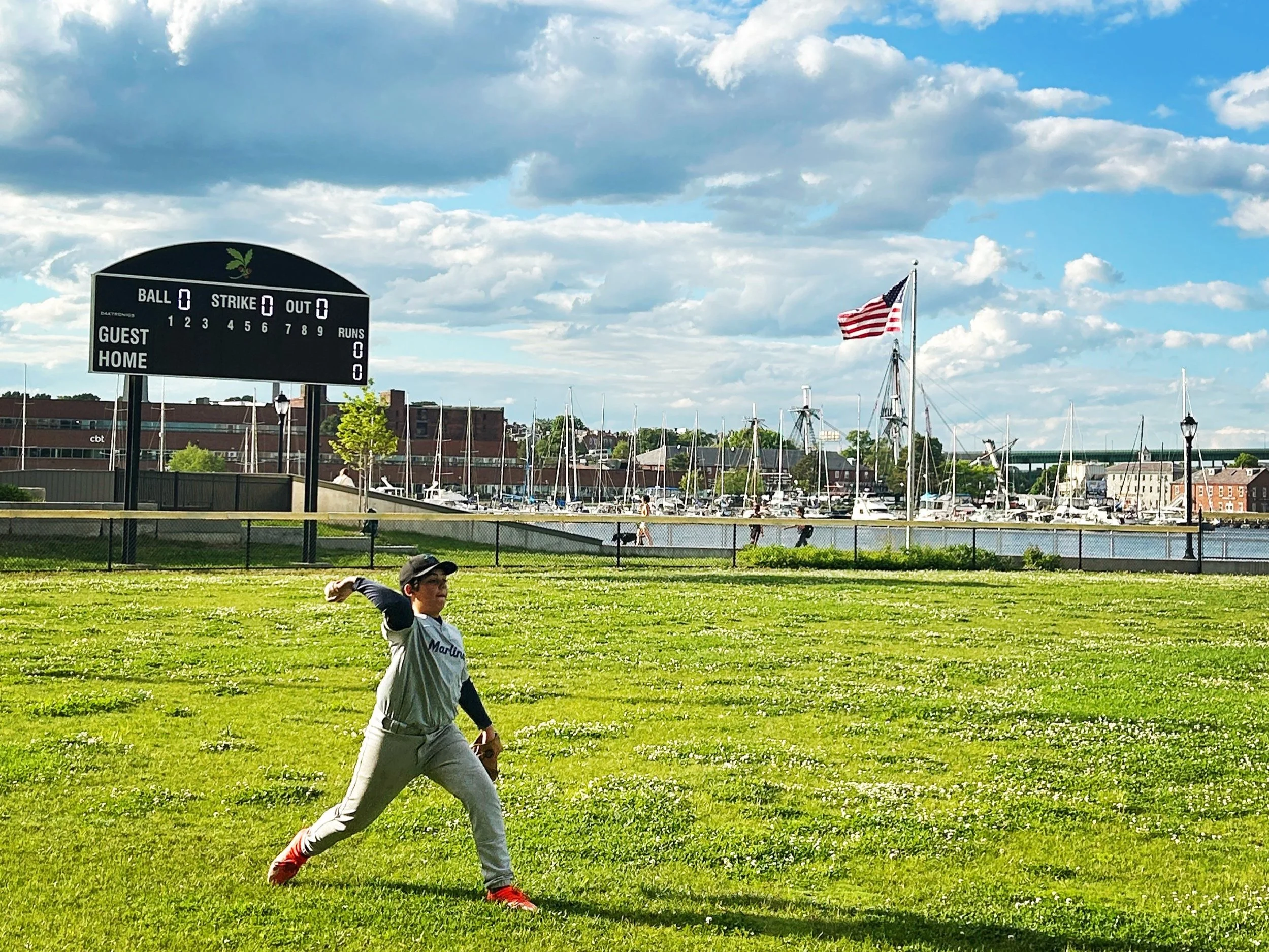 A young boy wearing a gray baseball jersey and red shoes is playing baseball on a grassy field near water with sailboats and an American flag in the background.