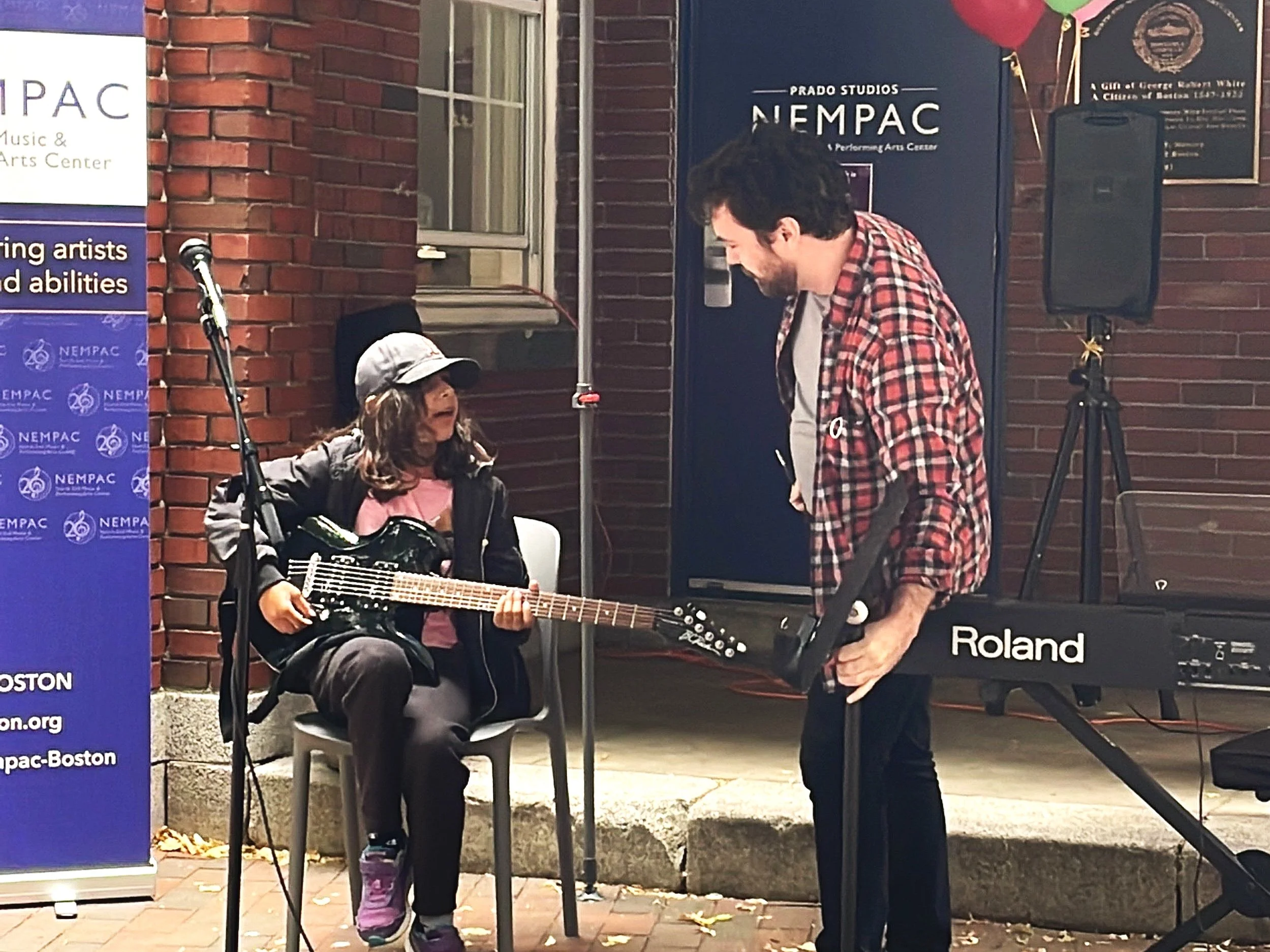A young girl with a guitar and a man with a keyboard engaging in music performance outside a brick building. The girl is sitting, singing or speaking to the man, who is standing and listening attentively. There is a blue banner with white text about NEMPAC on the left and musical equipment around them.