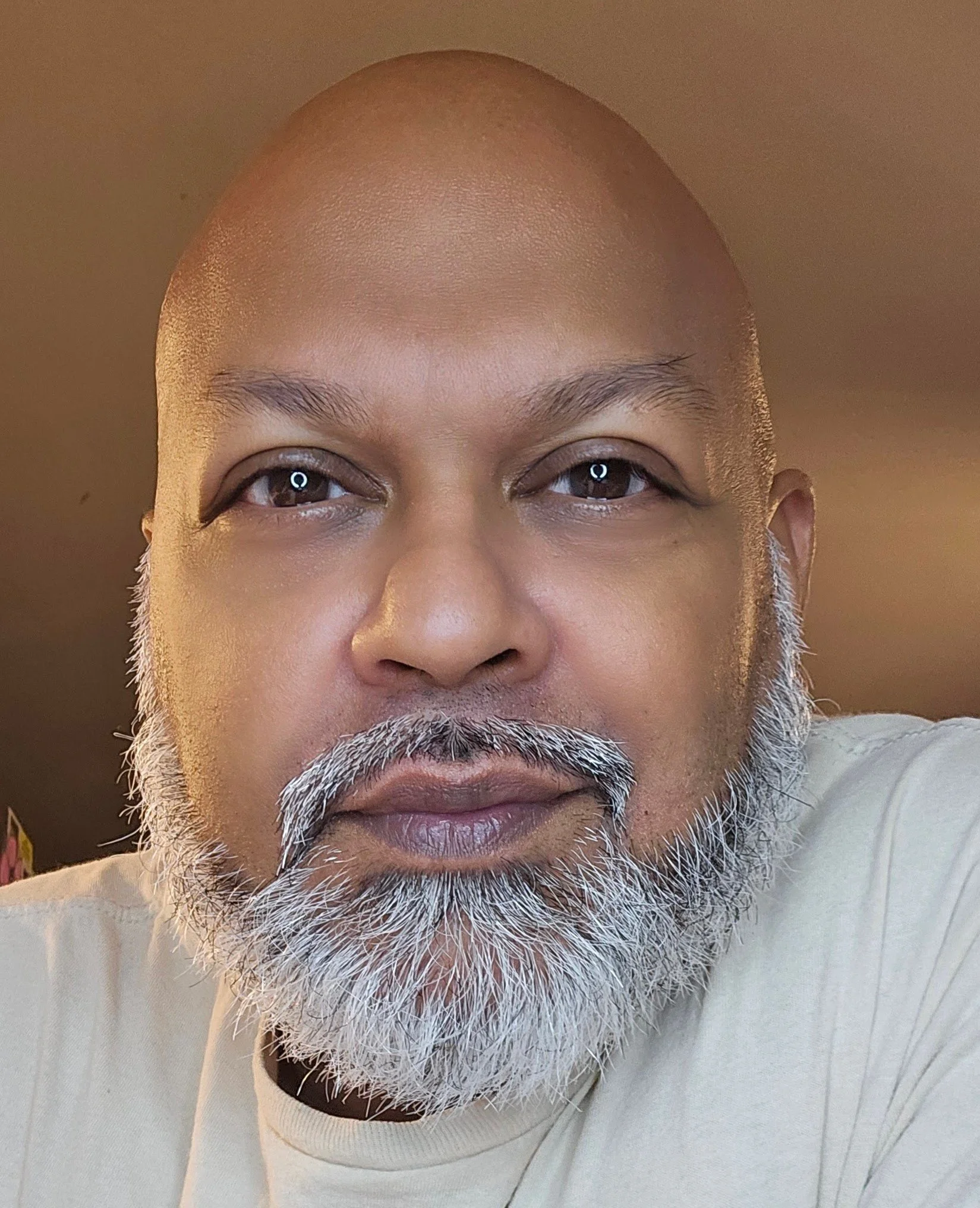Close-up of a man with a bald head, grey beard, and wearing a white shirt, looking into the camera with a neutral expression.