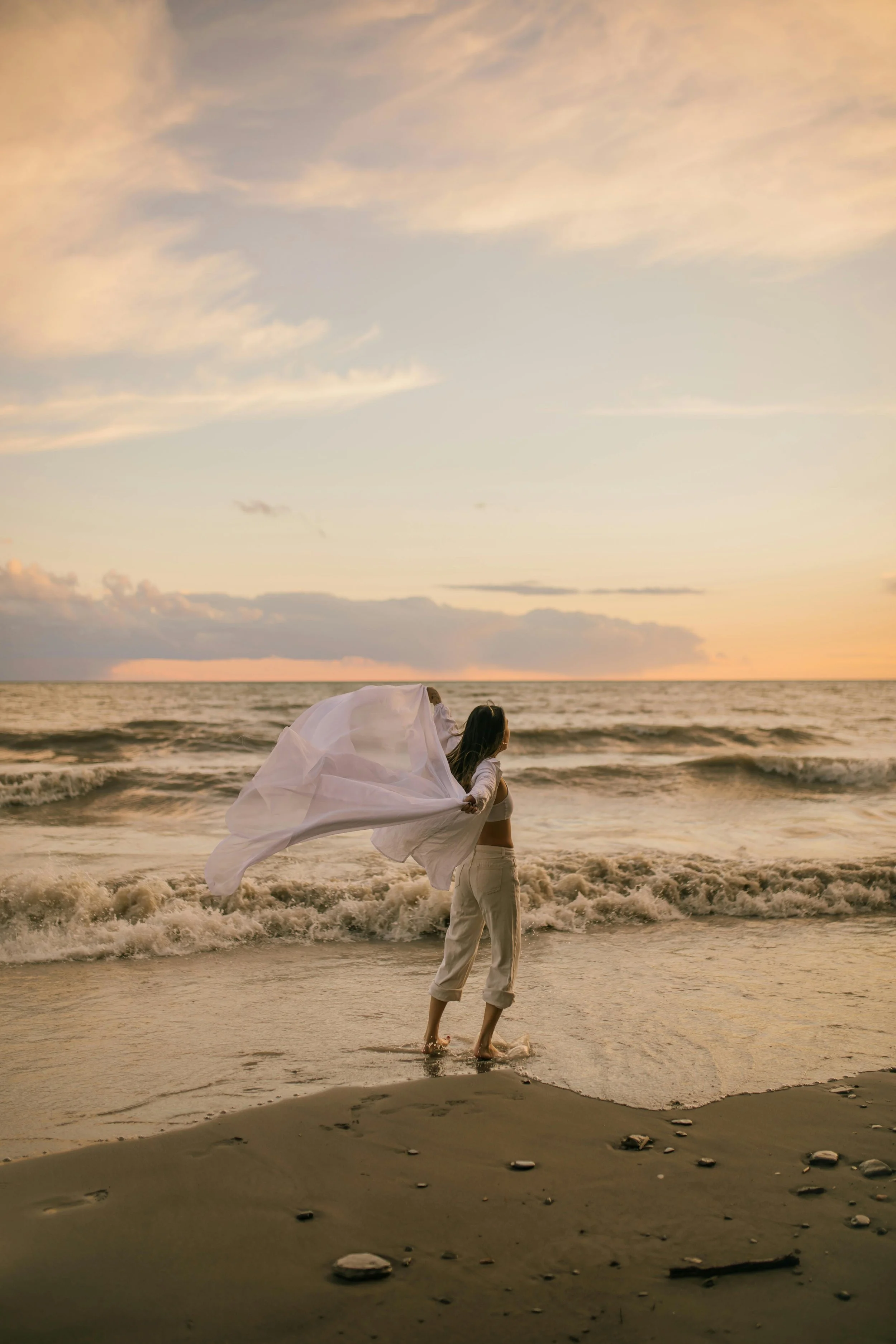 A woman holding a flowing white fabric on a beach at sunset.