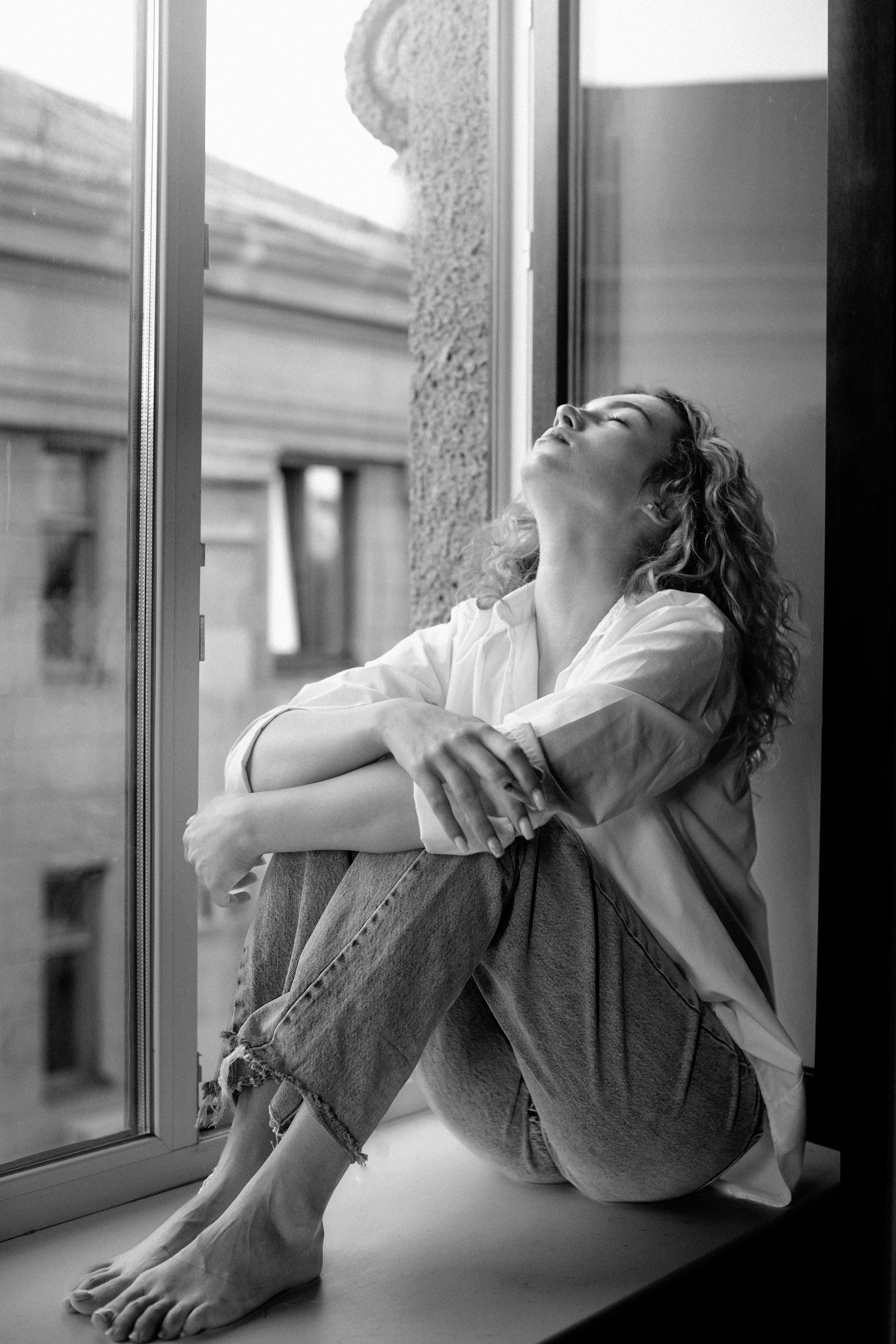 A woman sitting on a windowsill with her eyes closed, appearing relaxed. She has curly hair and is wearing a light-colored shirt and ripped jeans.