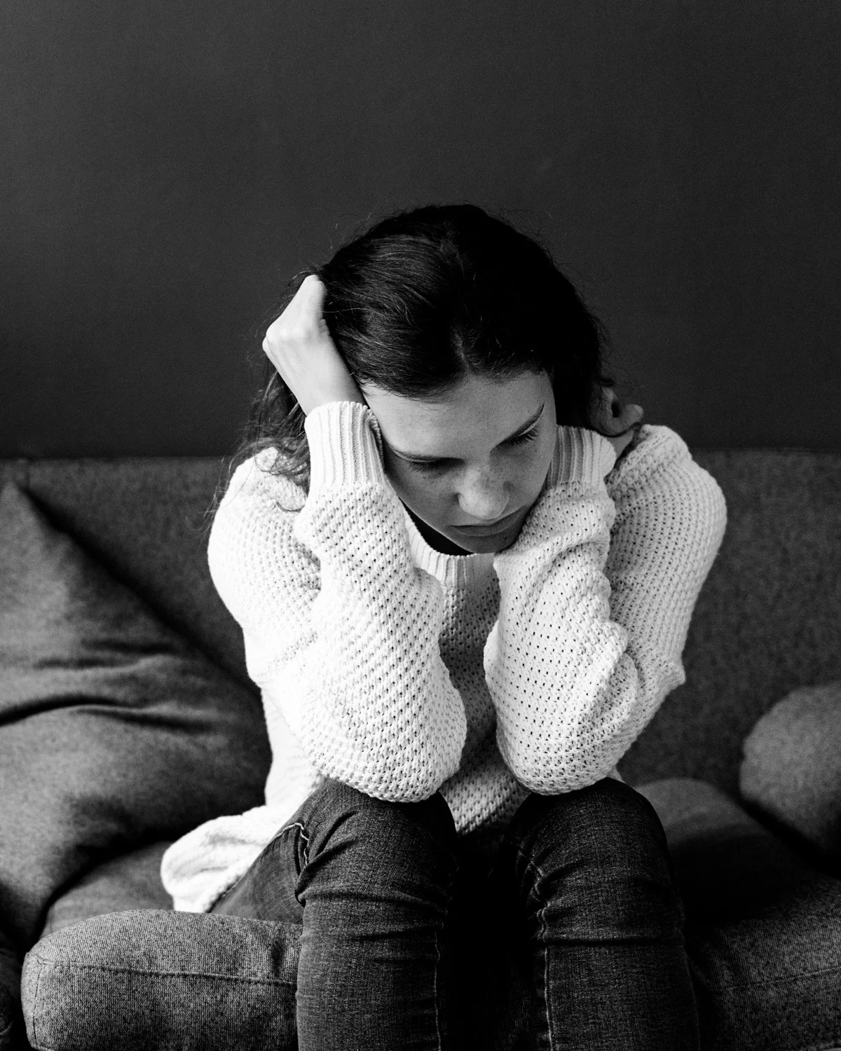 A woman with dark hair sitting on a couch, holding her head with both hands, looking downward with a serious or contemplative expression.