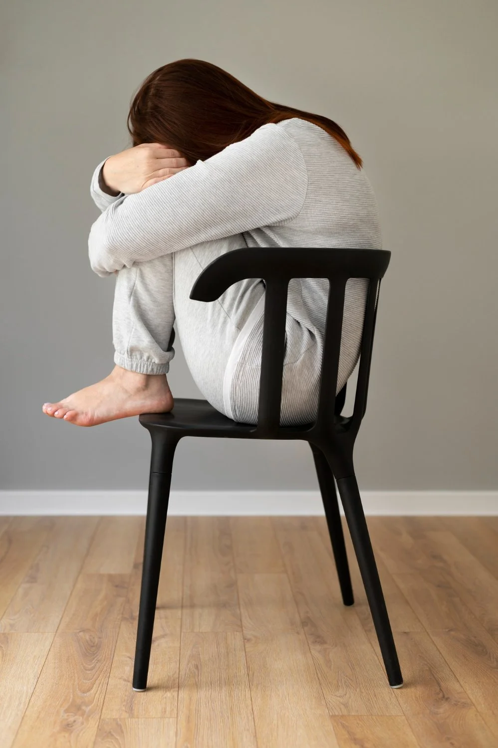 A woman with brown hair sitting on a black chair, hugging her knees to her chest, with her head resting on her arms. She is wearing a light grey outfit and is barefoot, with one foot resting on the chair and the other hanging down.
