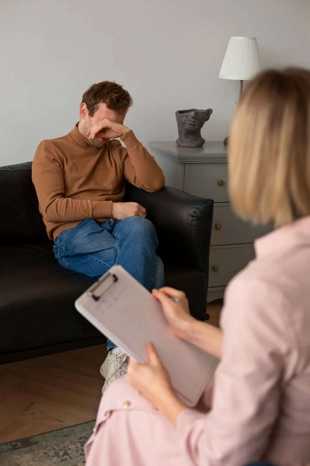A man sitting on a black sofa with his hand covering his face, appearing upset or distressed, while a woman holds a clipboard and a pen in front of him, interviewing or counseling him in a room with a lamp and decorative sculpture in the background.