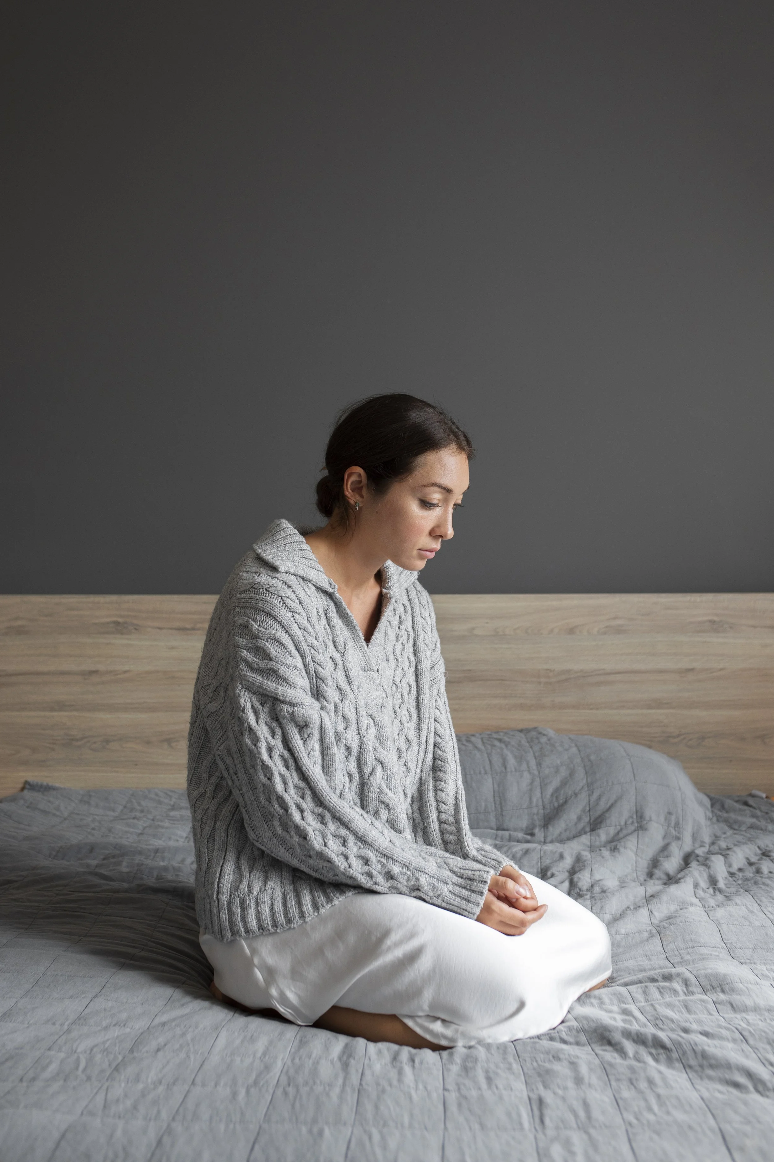 A woman sitting cross-legged on a bed with grey bedding, wearing a grey cable-knit sweater and white pants, in a room with a dark grey wall and wooden headboard.