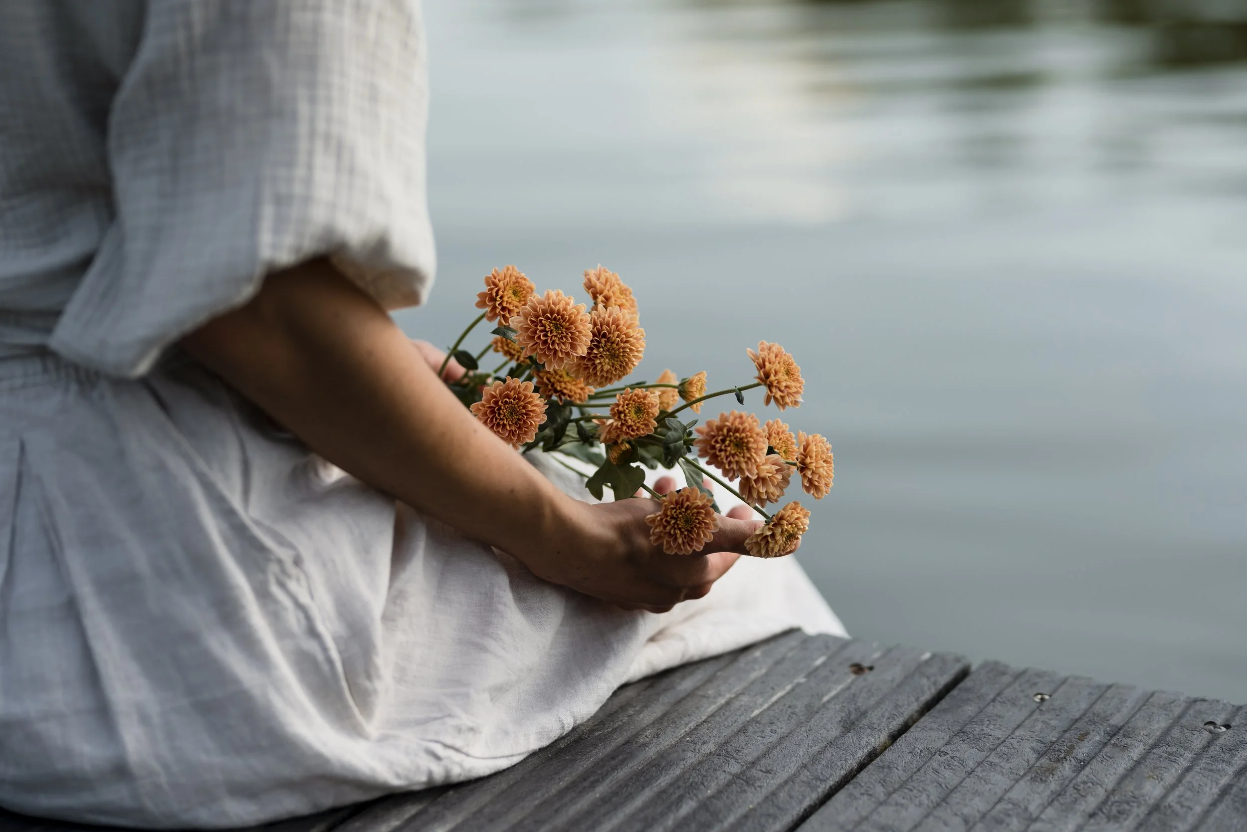 Person sitting on a wooden dock by a body of water, holding a bouquet of orange chrysanthemums.