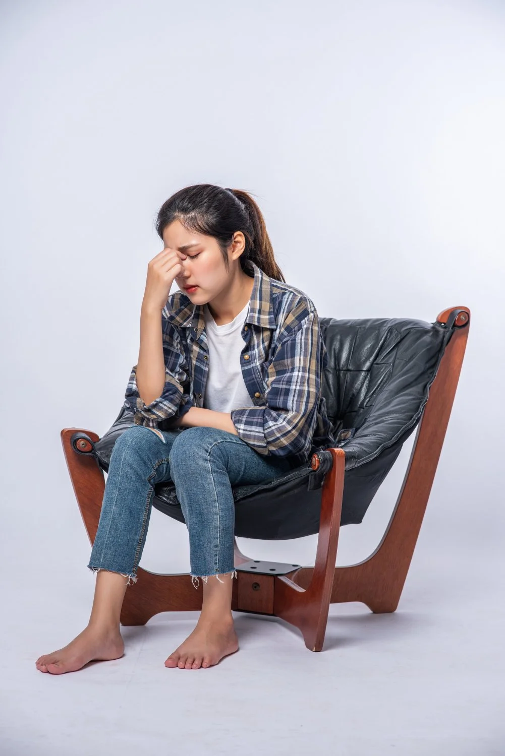 Young woman sitting on a black leather and wooden chair with her eyes closed and hand pinching the bridge of her nose, looking distressed, wearing a plaid shirt and jeans, with bare feet.