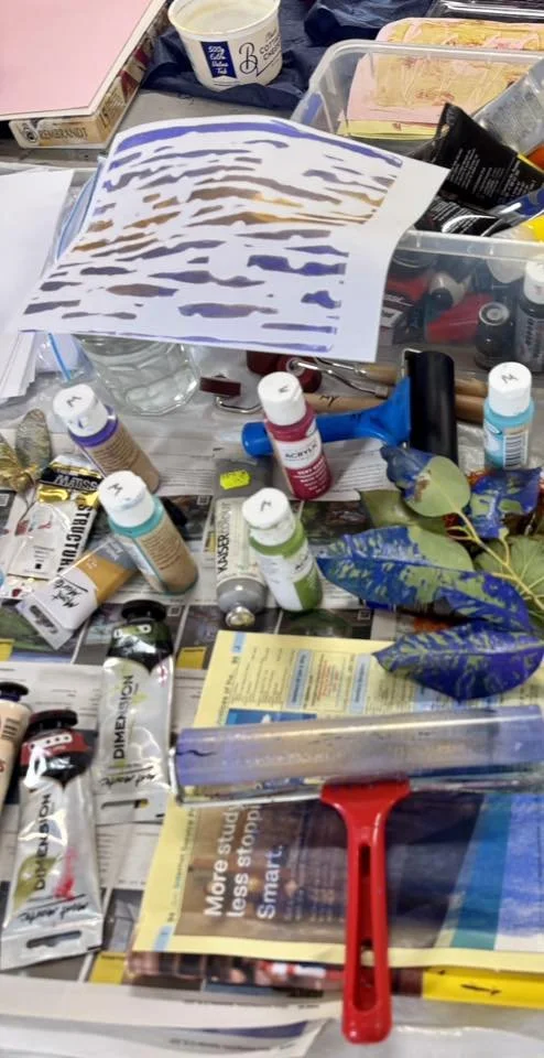 A cluttered workspace with paint bottles, a paint scraper, leaves, a plastic container with tubes, and various painting supplies.