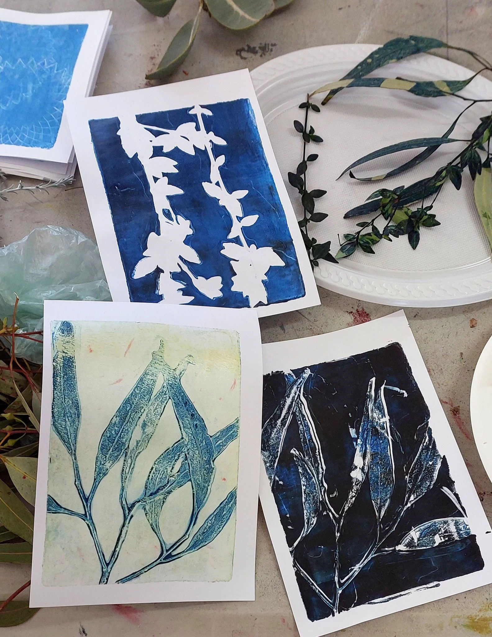 Three botanical print artworks with leaf patterns on white paper, displayed on a table along with some foliage and decorative plates.