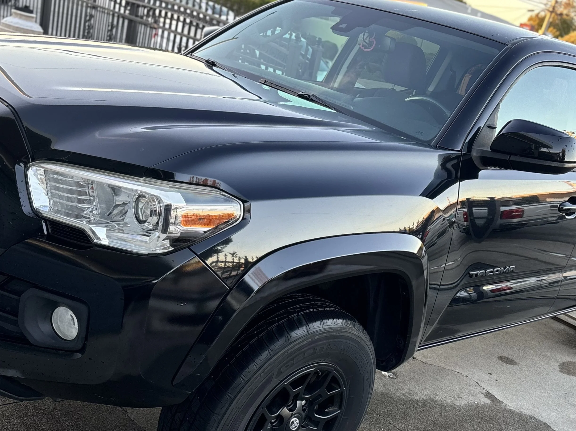 Front side of a black Toyota Tacoma pickup truck parked outdoors, showing headlight, front wheel, and part of the windshield.