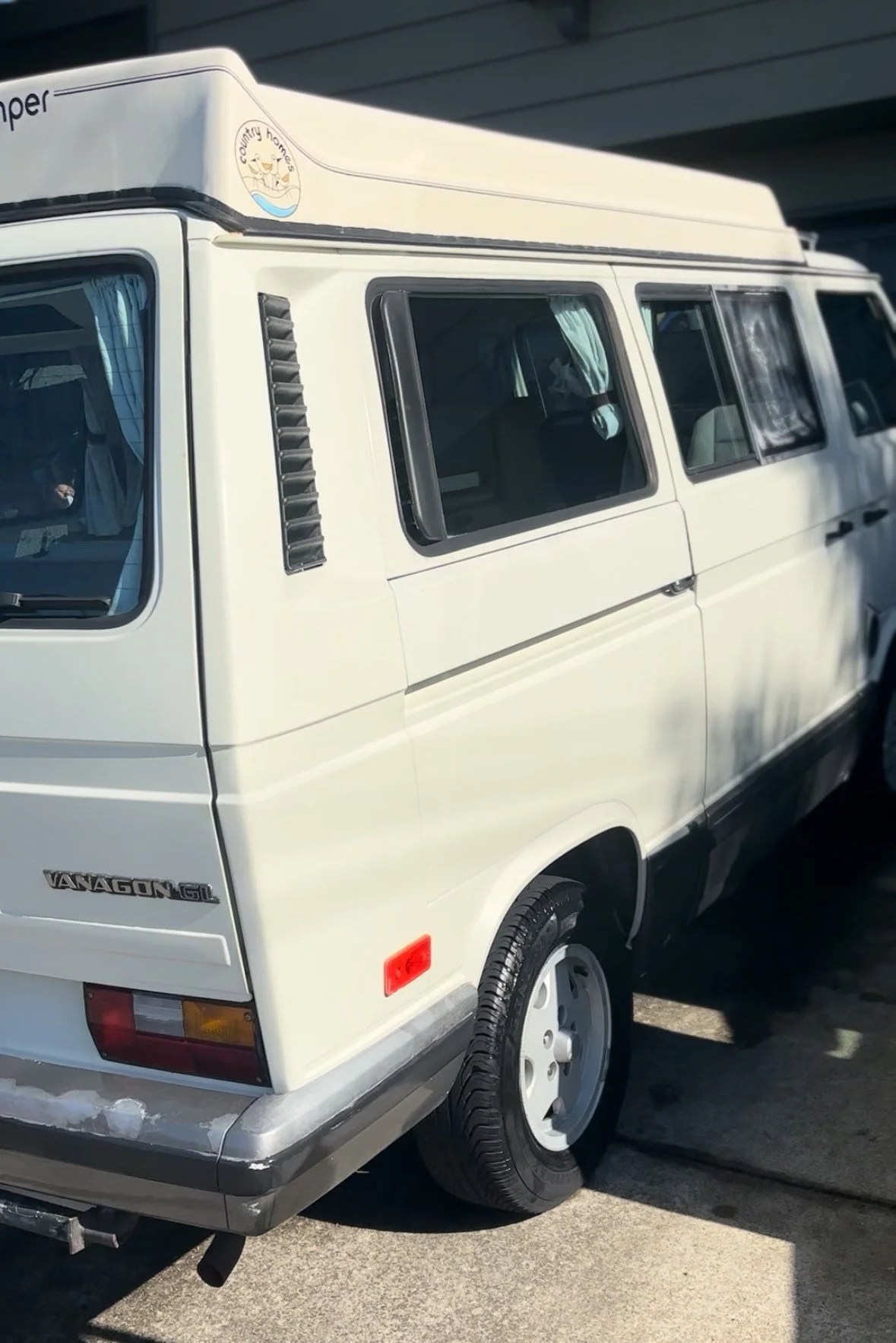 White Volkswagen Vanagon GL parked outdoors with tinted windows, black vent slats, and a decal on the roof that says "country homes."